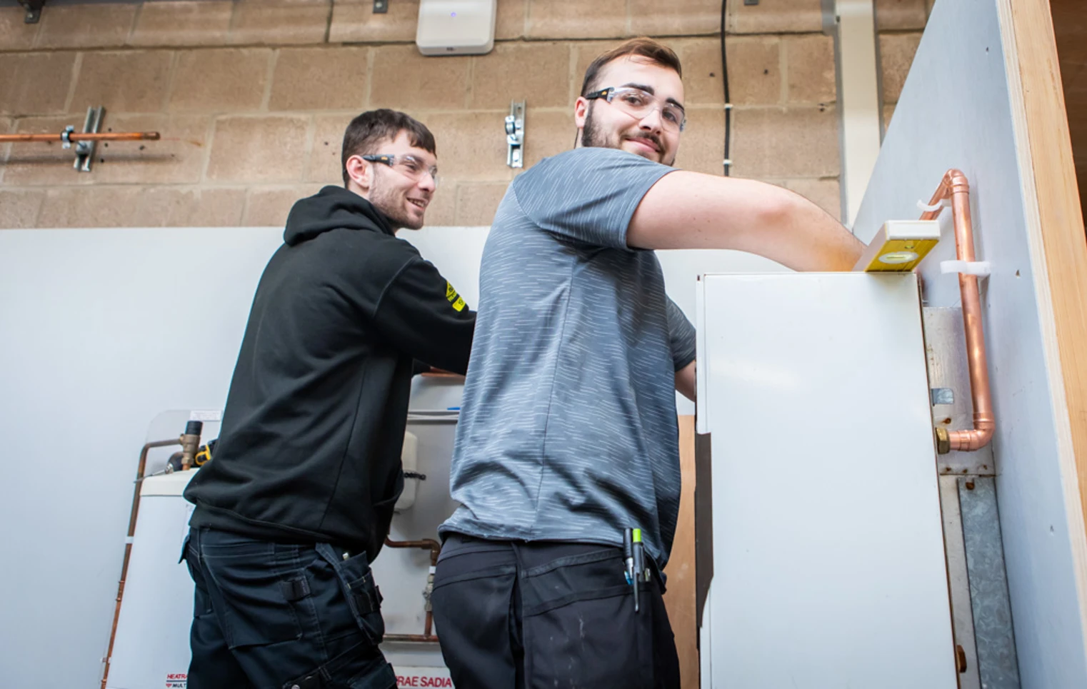 Two plumbing students smiling while collaboratively working on installing a plumbing fixture, engaged in hands-on training. Two plumbing students smiling while collaboratively working on installing a plumbing fixture, engaged in hands-on training.