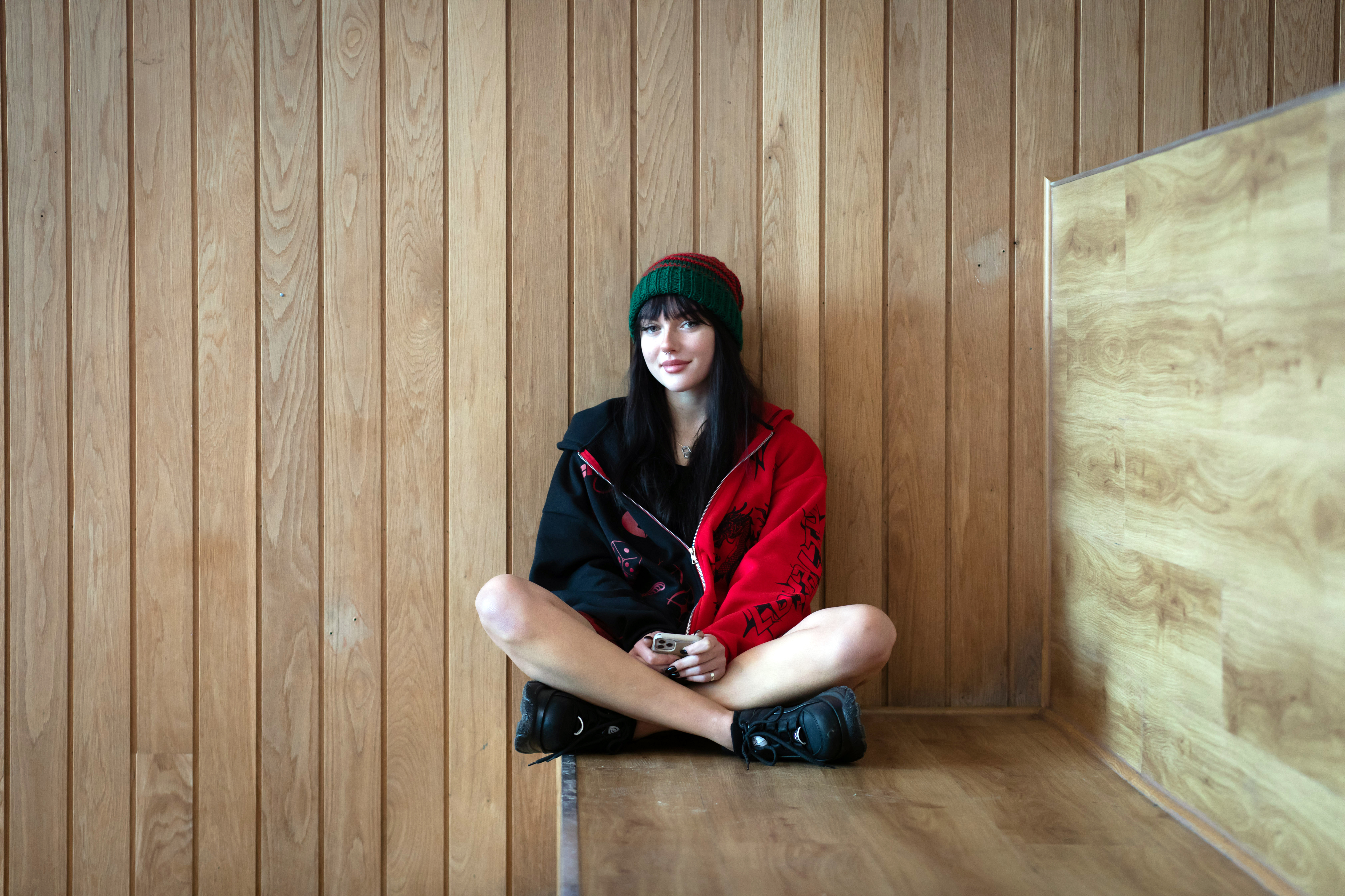 Student sitting cross-legged on a wooden bench against a wood-panelled wall at Glasgow Kelvin College’s Springburn Campus, wearing a red and black jacket and green hat.
