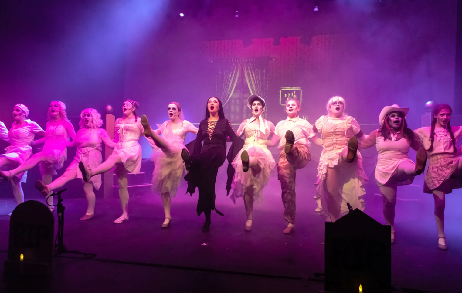 Stage performance featuring a line of dancers in ghostly white outfits, led by a character in dark clothing, all kicking in perfect unison. Stage performance featuring a line of dancers in ghostly white outfits, led by a character in dark clothing, all kicking in perfect unison.
