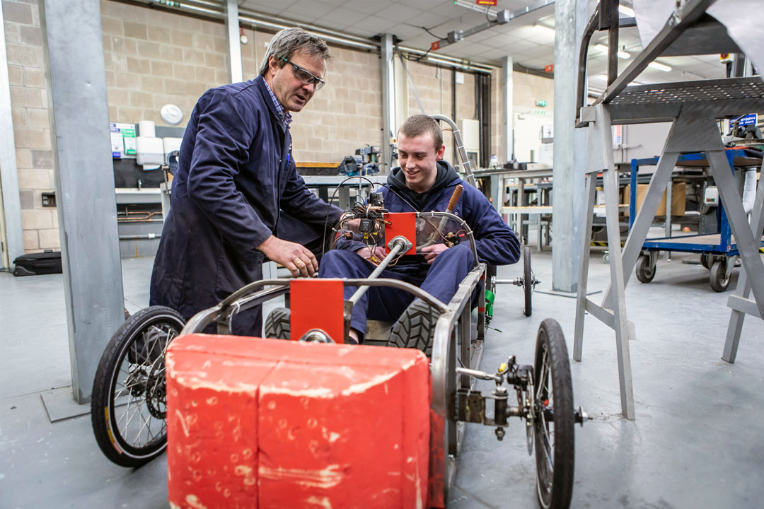 A lecturer assisting a student seated in a custom-built electric car, adjusting the wiring and components during a hands-on engineering project.