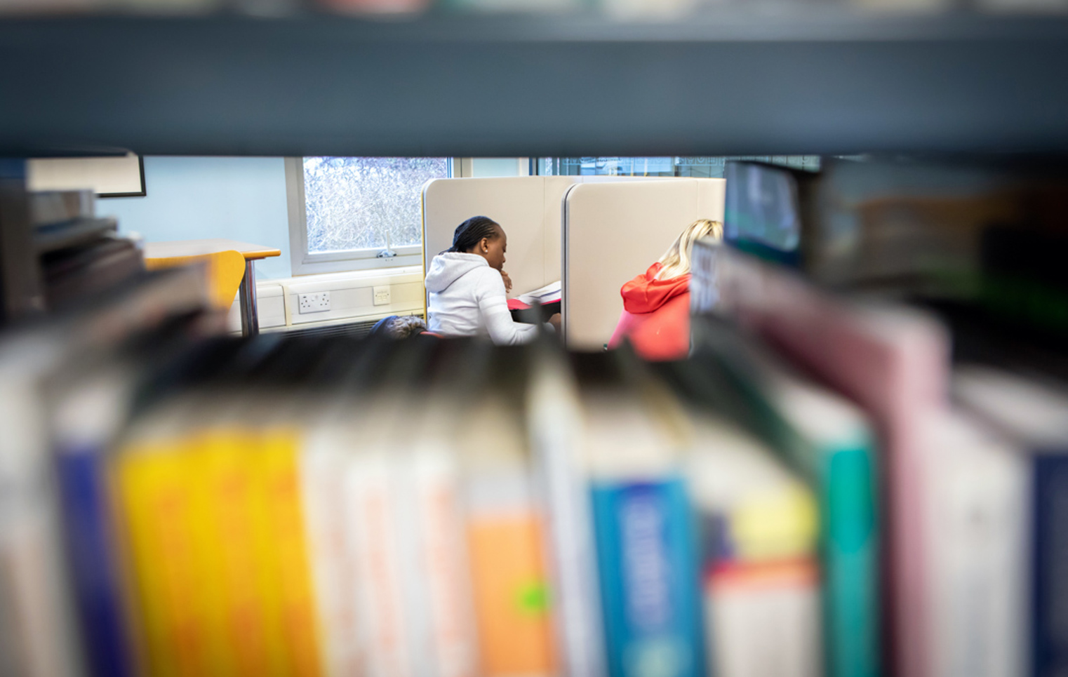 Students studying in individual booths at the East End campus library, viewed through a shelf of books.