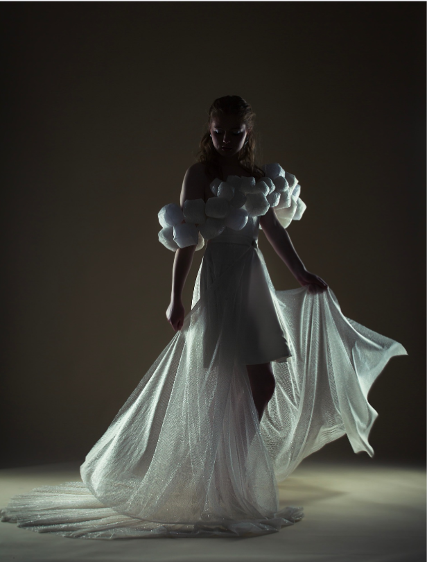 Model in a dramatic, low-lit setting wearing an off-the-shoulder white gown with textured puff sleeves and a flowing sheer train, softly illuminated from behind.