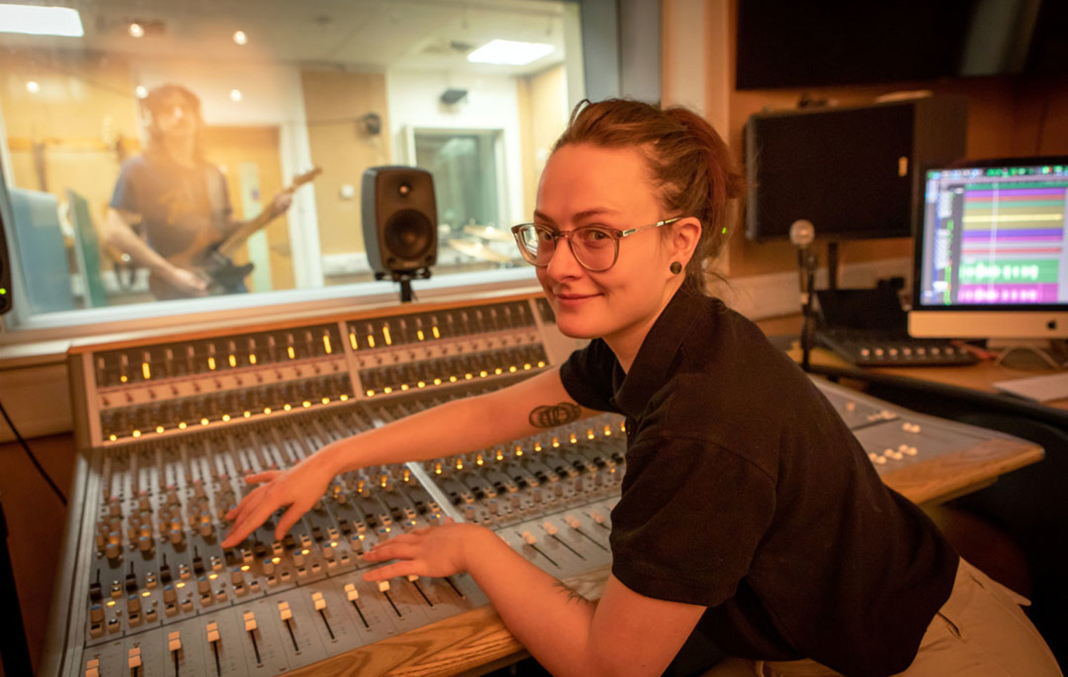 A student in a black polo shirt adjusting the controls on a recording studio mixing desk while smiling at the camera.