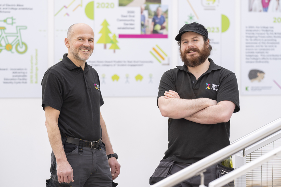 Two male staff members from the Glasgow Kelvin College Estates Team standing indoors, smiling at the camera in front of sustainability-themed posters.
