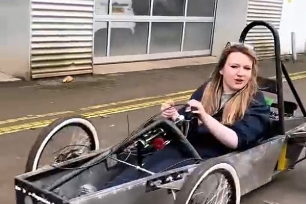 Automotive student driving a student-built race car during a practical session outside the workshop.