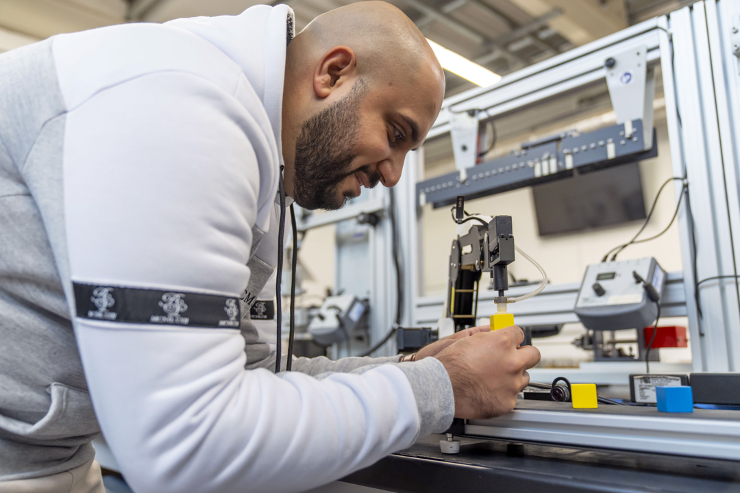 Student operating a robotic arm, carefully placing a yellow block on a conveyor system as part of an automation project