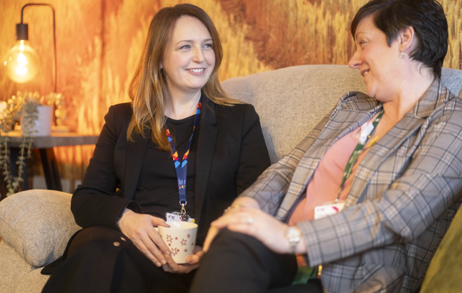Two Glasgow Kelvin College staff members sitting on a sofa, smiling and chatting in a relaxed setting with warm lighting Two Glasgow Kelvin College staff members sitting on a sofa, smiling and chatting in a relaxed setting with warm lighting