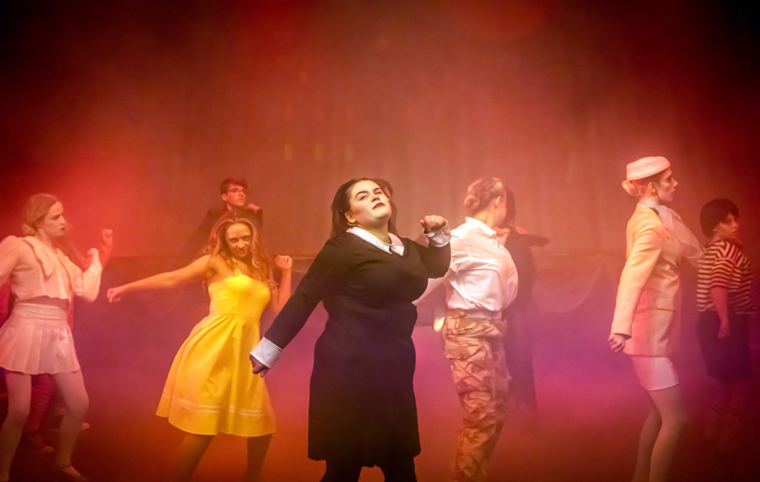 Musical theatre students dancing in formation during a stage performance, wearing contrasting costumes and performing under warm, atmospheric lighting.