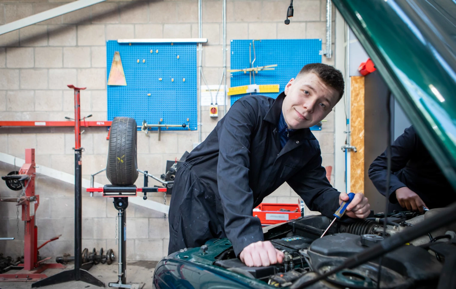 Student in a black overall smiling while working on a car engine, with a workshop setup in the background. Student in a black overall smiling while working on a car engine, with a workshop setup in the background.