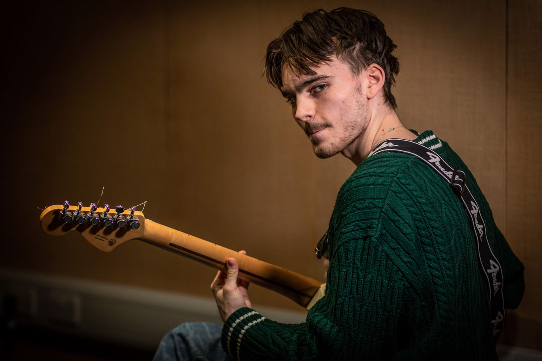 A young guitarist wearing a green sweater, holding a Fender guitar, and looking towards the camera in a dimly lit practice room.