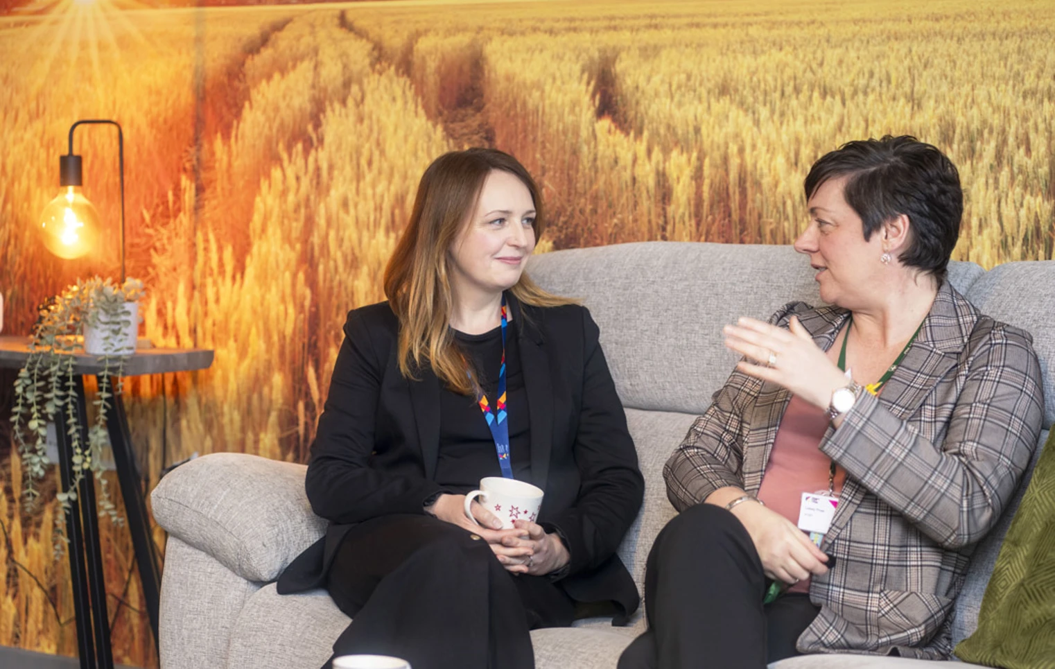 Two Glasgow Kelvin College staff members sitting on a sofa with coffee mugs, having a friendly conversation in a relaxed setting with a scenic wall backdrop. Two Glasgow Kelvin College staff members sitting on a sofa with coffee mugs, having a friendly conversation in a relaxed setting with a scenic wall backdrop.