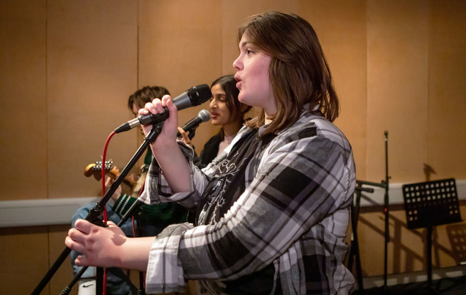 A close-up of a young singer holding a microphone and performing with a student band in a rehearsal room.
