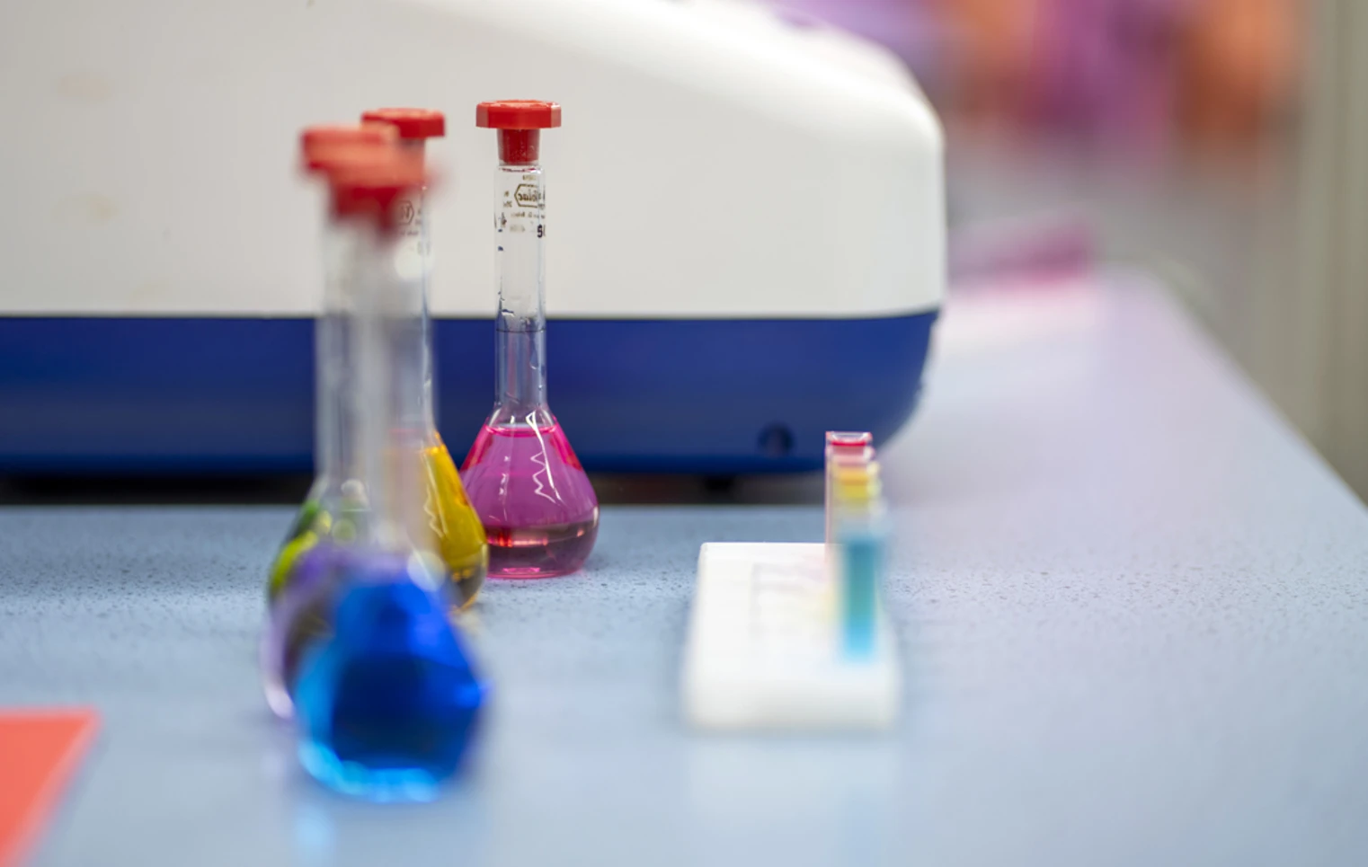 Three glass flasks filled with bright blue, yellow, and pink liquids sit on a lab bench, with a row of colourful test tubes and a science machine in the background—ready for a splash of science fun and discovery. Three glass flasks filled with bright blue, yellow, and pink liquids sit on a lab bench, with a row of colourful test tubes and a science machine in the background—ready for a splash of science fun and discovery.