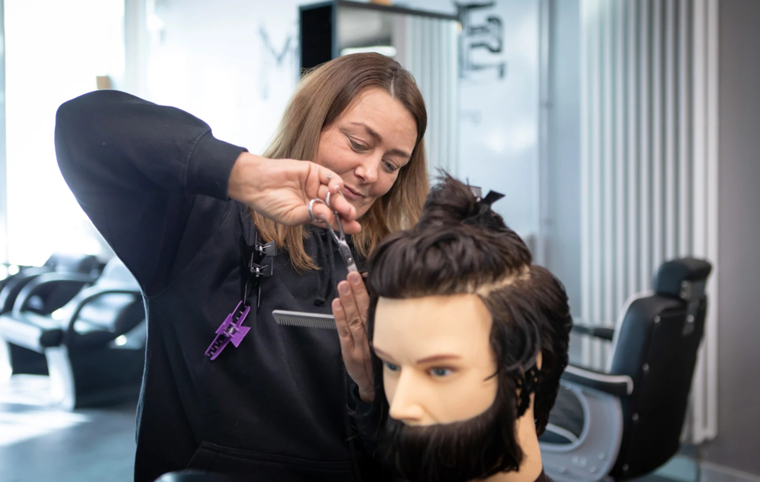 A female barbering student in a black hoodie carefully cutting the hair on a mannequin head inside a training facility. A female barbering student in a black hoodie carefully cutting the hair on a mannequin head inside a training facility.