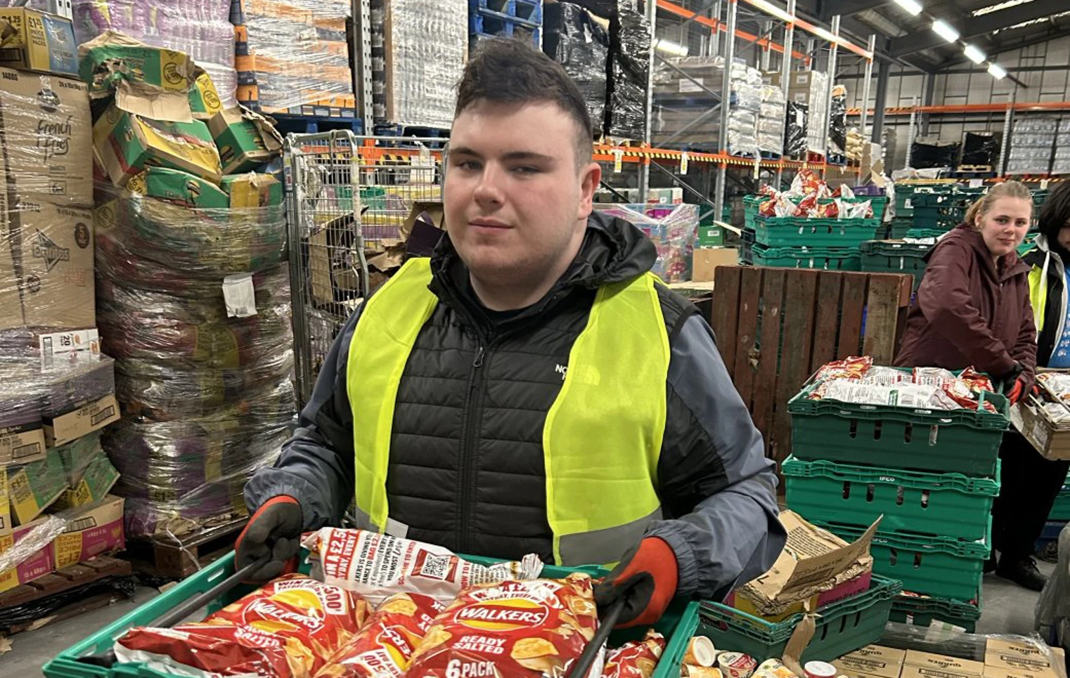 Young man wearing a high-visibility vest holding a crate of packaged crisps while working in a warehouse environment. Young man wearing a high-visibility vest holding a crate of packaged crisps while working in a warehouse environment.