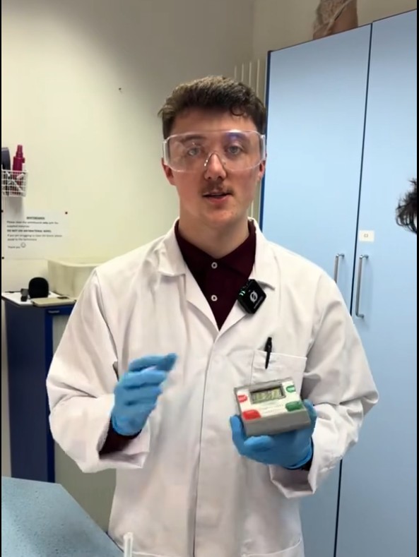 Science student Lewis wearing safety goggles and gloves while demonstrating lab equipment in a college laboratory.