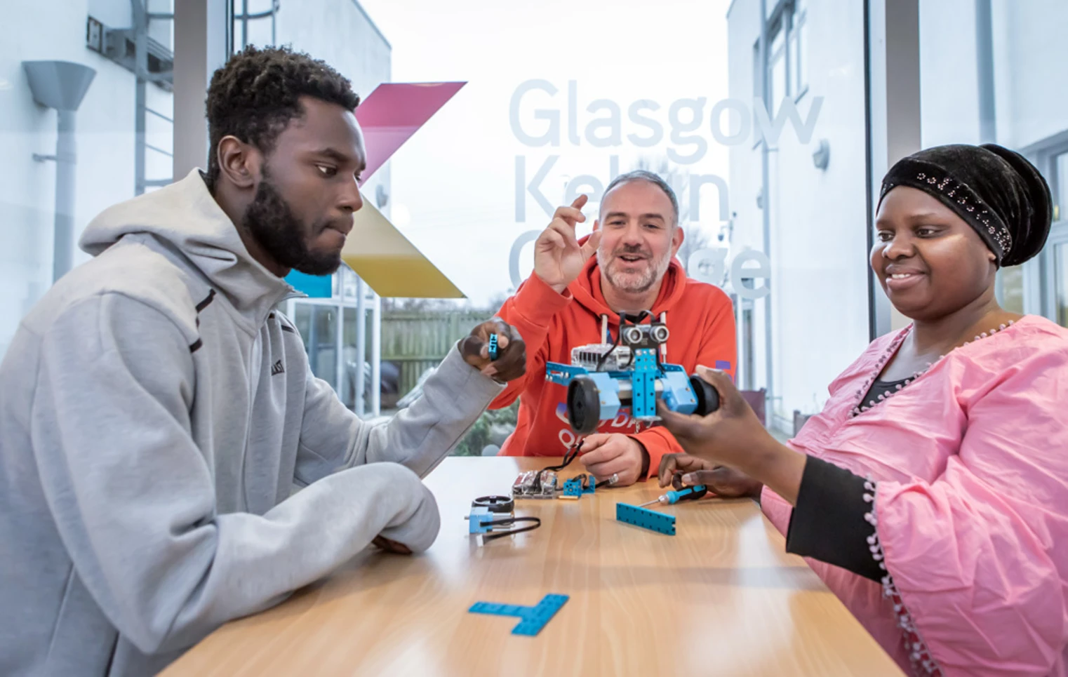Lecturer in an orange hoodie helping two students troubleshoot and assemble a blue robotic vehicle in a classroom. Lecturer in an orange hoodie helping two students troubleshoot and assemble a blue robotic vehicle in a classroom.