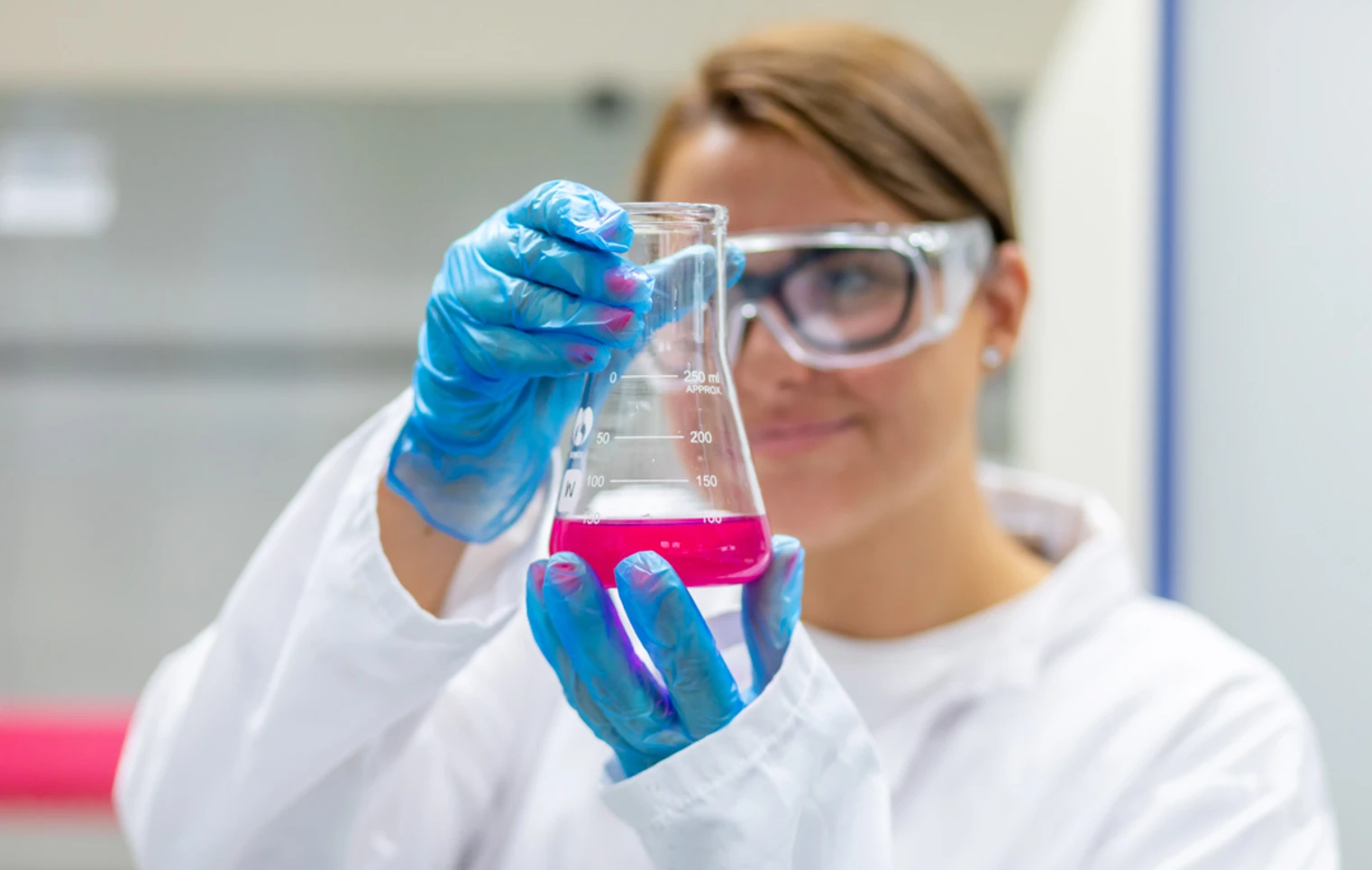 Student holding a beaker of pink liquid close to the camera, wearing gloves and safety goggles. Student holding a beaker of pink liquid close to the camera, wearing gloves and safety goggles.