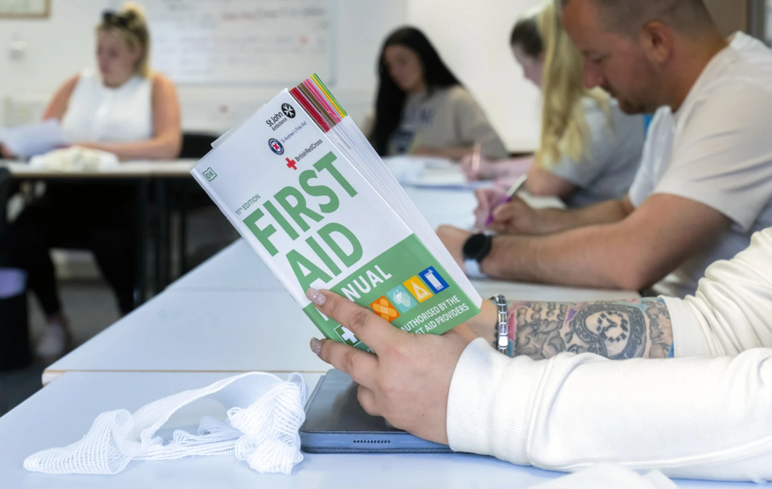 A close-up of a tattooed hand holding a first aid manual above a desk, with a white bandage and a tablet nearby—serious about learning, with a bit of style A close-up of a tattooed hand holding a first aid manual above a desk, with a white bandage and a tablet nearby—serious about learning, with a bit of style