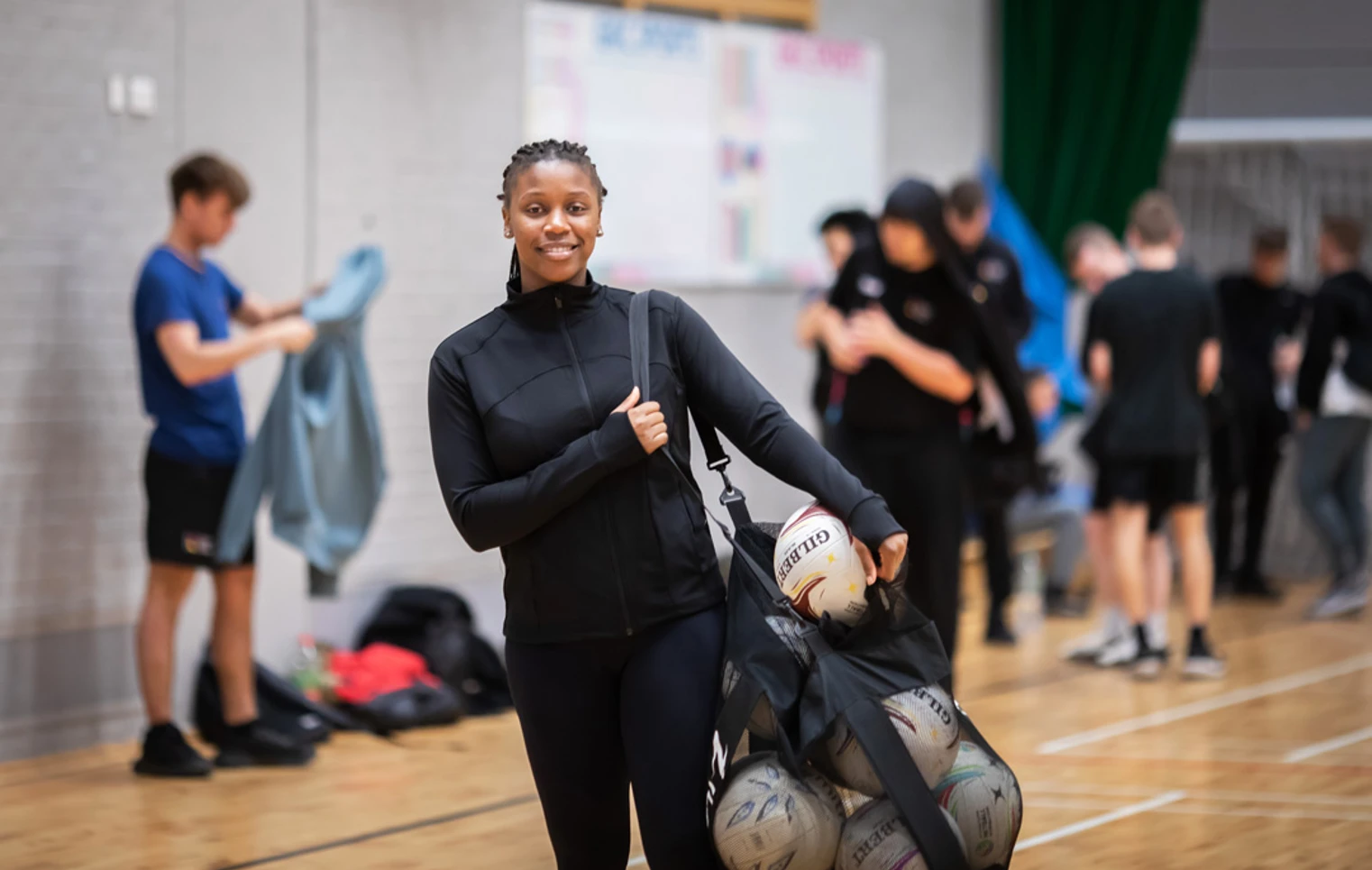 A student holding a mesh bag filled with netballs, smiling at the camera in a sports hall. A student holding a mesh bag filled with netballs, smiling at the camera in a sports hall.