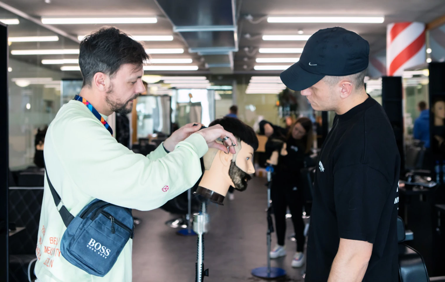 A barbering instructor demonstrating a haircut on a mannequin head while a student in a black cap observes. A barbering instructor demonstrating a haircut on a mannequin head while a student in a black cap observes.