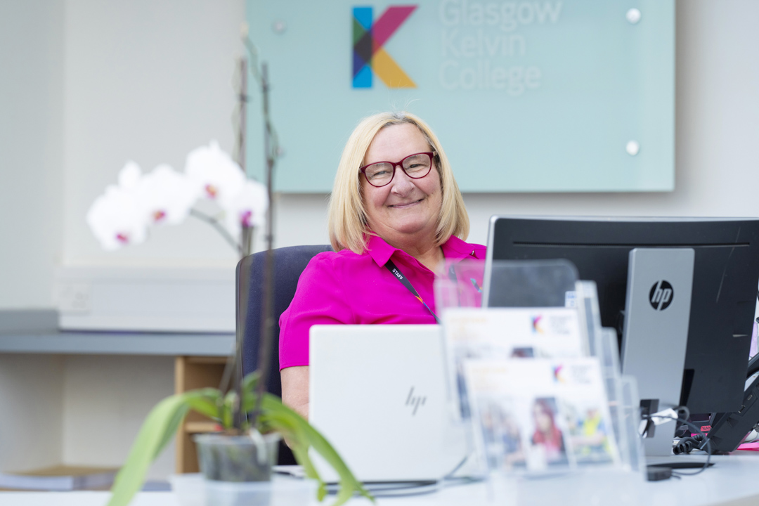 Receptionist smiling at her desk in the Glasgow Kelvin College West Campus, with a Glasgow Kelvin College sign in the background and orchids in the foreground.