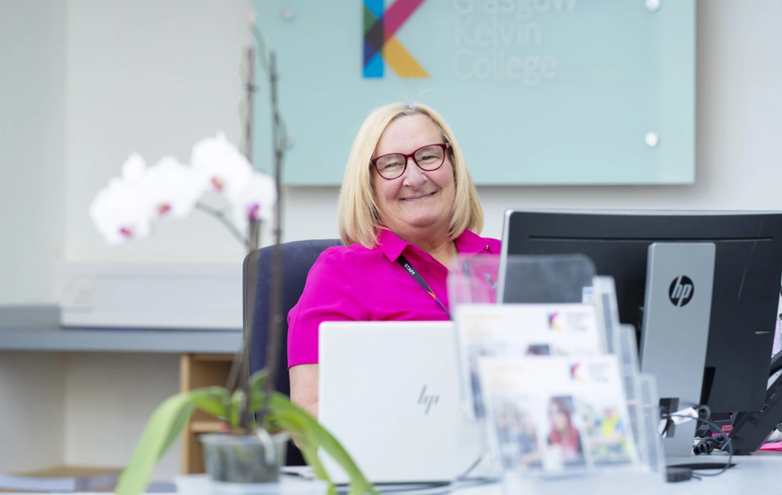 Receptionist smiling at her desk in the Glasgow Kelvin College West Campus, with a Glasgow Kelvin College sign in the background and orchids in the foreground. Receptionist smiling at her desk in the Glasgow Kelvin College West Campus, with a Glasgow Kelvin College sign in the background and orchids in the foreground.