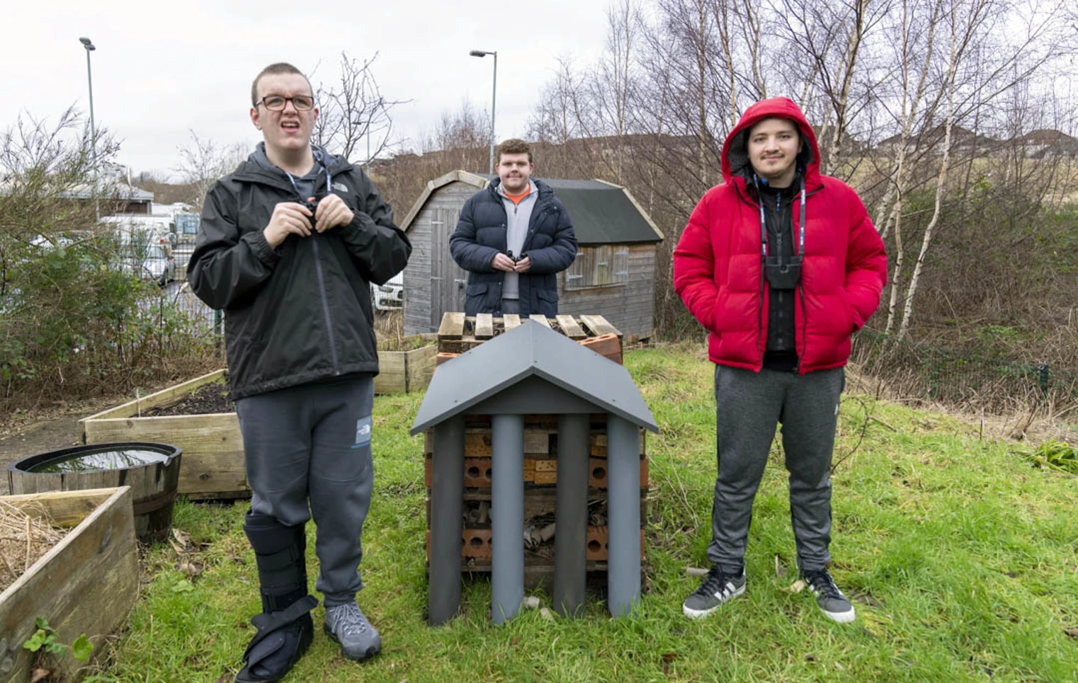 Three young men standing outside beside a handmade bug hotel, surrounded by greenery and raised garden beds. Three young men standing outside beside a handmade bug hotel, surrounded by greenery and raised garden beds.