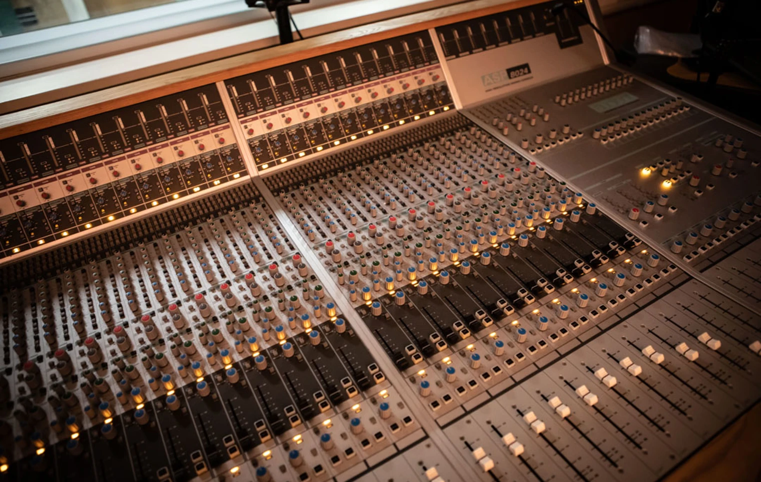 A wide shot of a professional recording studio mixing desk, with multiple sliders, knobs, and glowing lights. A wide shot of a professional recording studio mixing desk, with multiple sliders, knobs, and glowing lights.