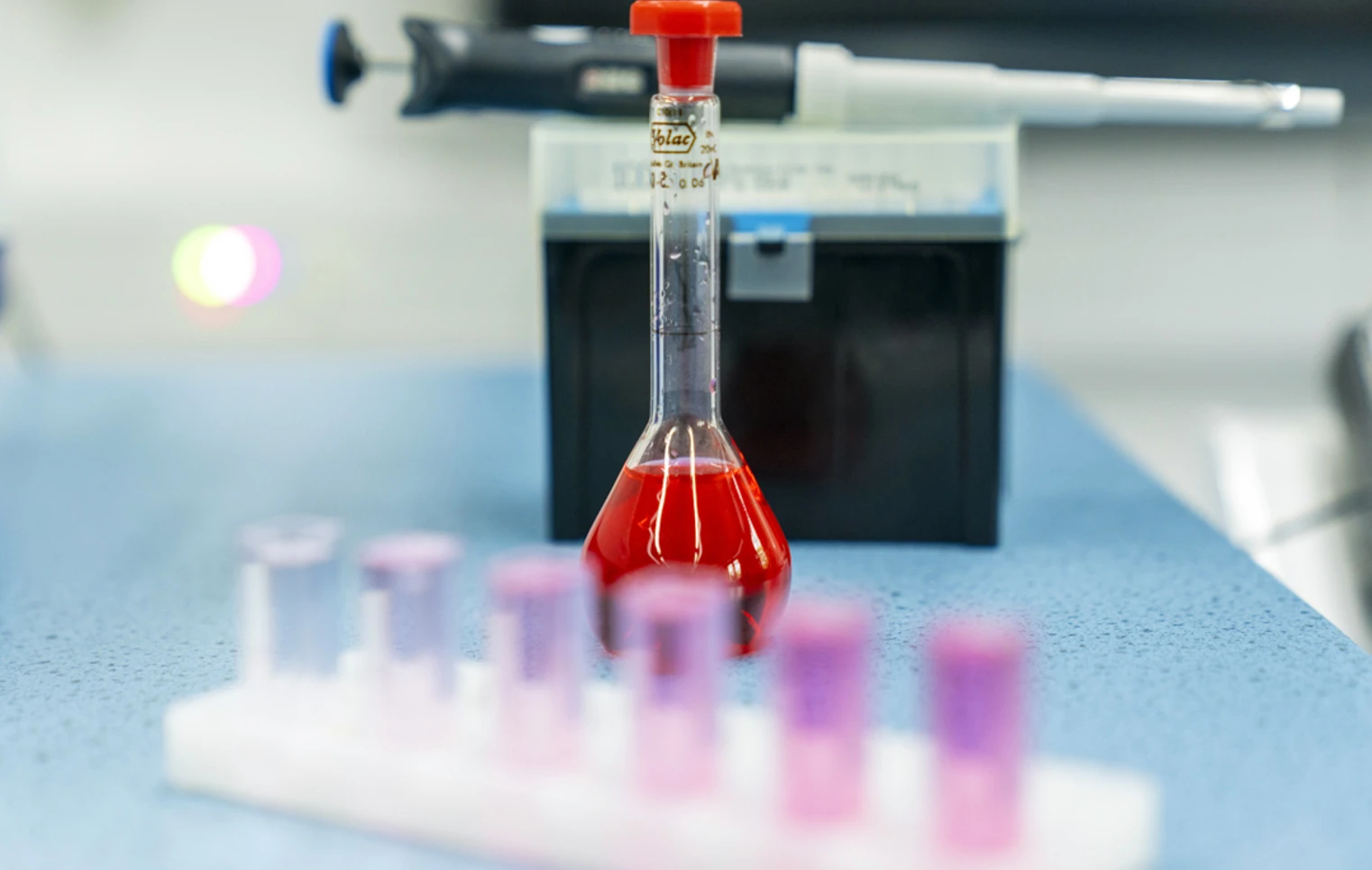 A bright red liquid fills a glass flask on a blue lab bench, with a row of test tubes in soft pinks and purples in front and a pipette resting in the background—science in action and full of colour. A bright red liquid fills a glass flask on a blue lab bench, with a row of test tubes in soft pinks and purples in front and a pipette resting in the background—science in action and full of colour.
