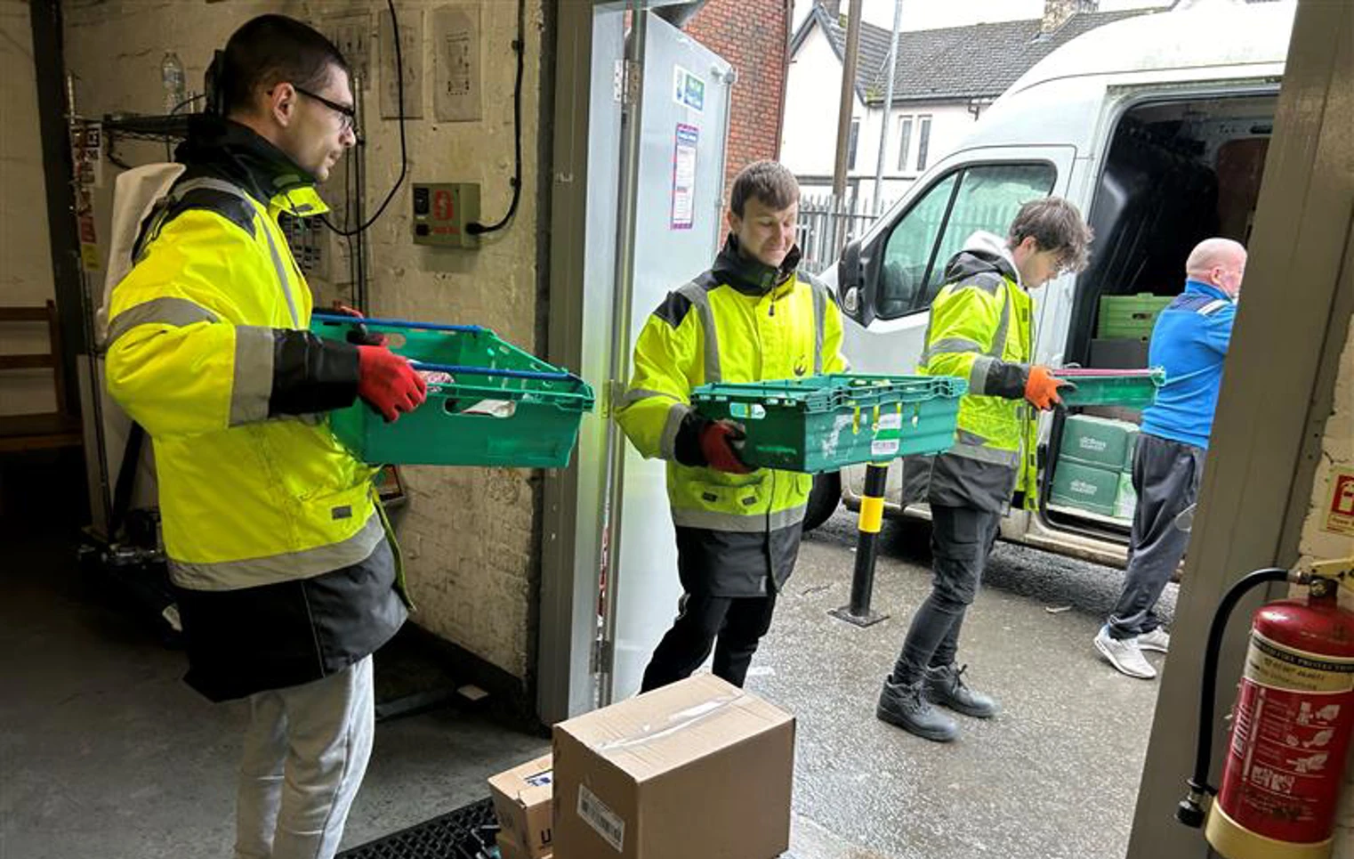 Three young men in high-visibility jackets unloading green crates from a delivery van at a warehouse entrance. Three young men in high-visibility jackets unloading green crates from a delivery van at a warehouse entrance.