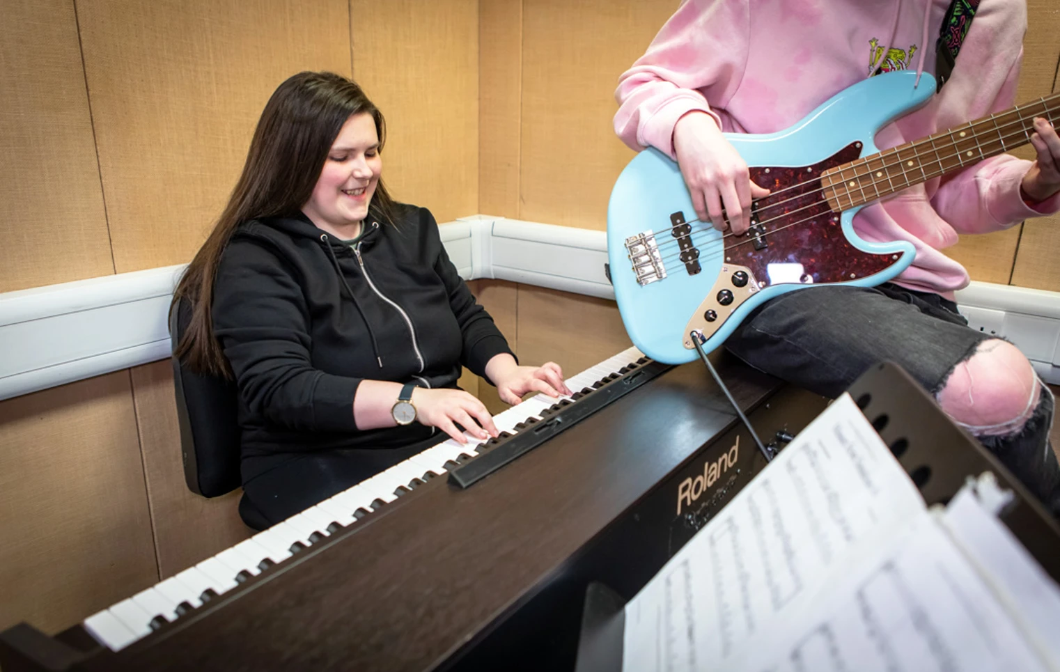 A smiling student in a black hoodie plays a Roland keyboard while another plays a blue bass guitar beside them. A smiling student in a black hoodie plays a Roland keyboard while another plays a blue bass guitar beside them.