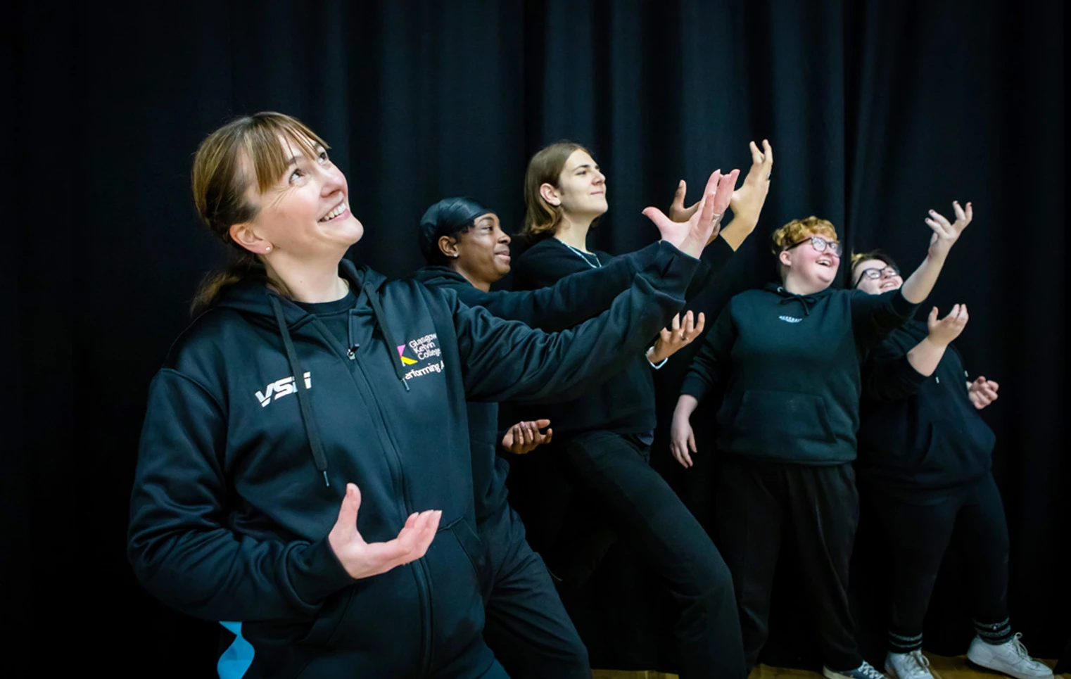 A lecturer and students striking dramatic poses against a black curtain, smiling and enjoying their creative acting activity. A lecturer and students striking dramatic poses against a black curtain, smiling and enjoying their creative acting activity.