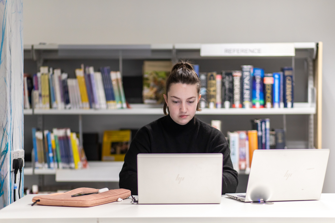 Student working on a laptop in the library at Glasgow Kelvin College, seated at a desk with bookshelves in the background.