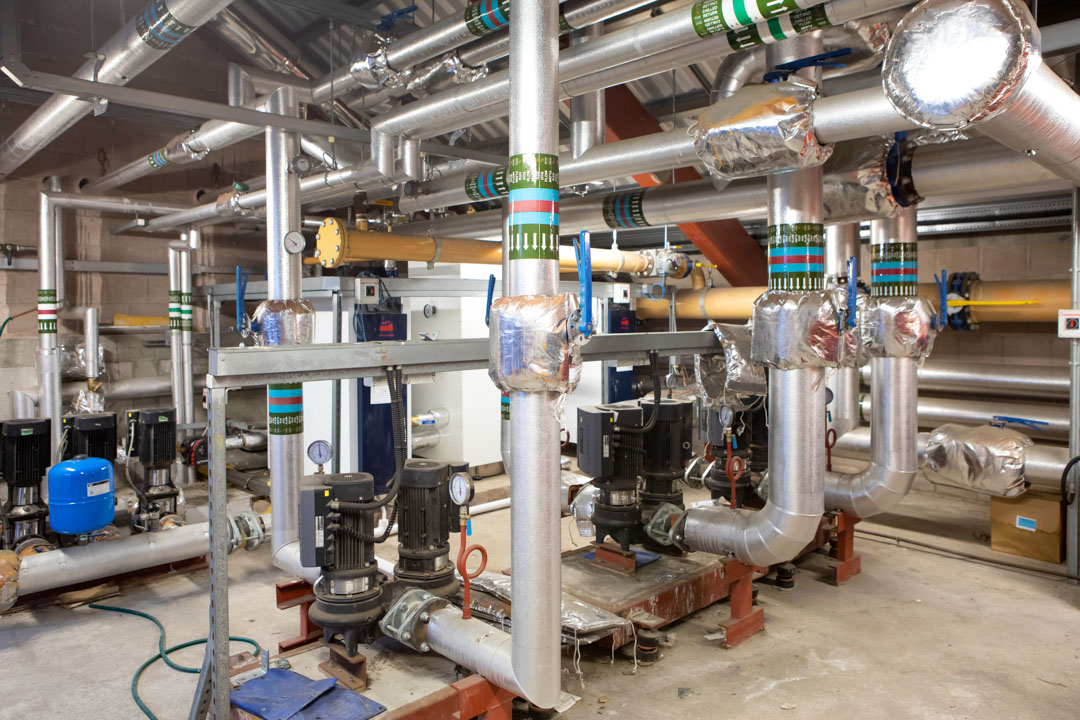 A network of insulated industrial pipes, pumps, and control valves in a large building services training facility