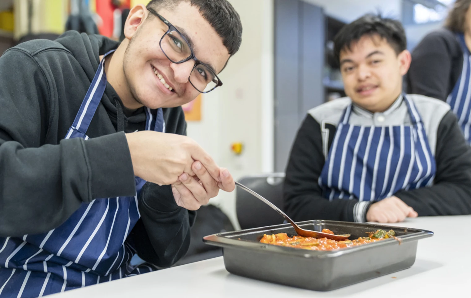 Two young men smiling and preparing a tray of food, wearing striped aprons in a kitchen setting. Two young men smiling and preparing a tray of food, wearing striped aprons in a kitchen setting.
