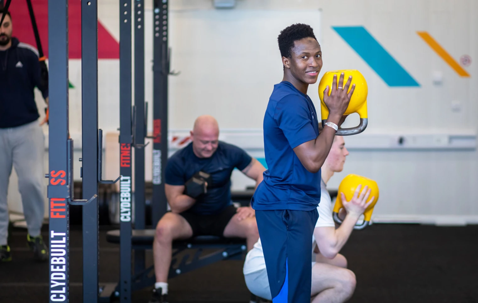 Student smiling while holding a yellow kettlebell during a strength training session at the gym. Student smiling while holding a yellow kettlebell during a strength training session at the gym.