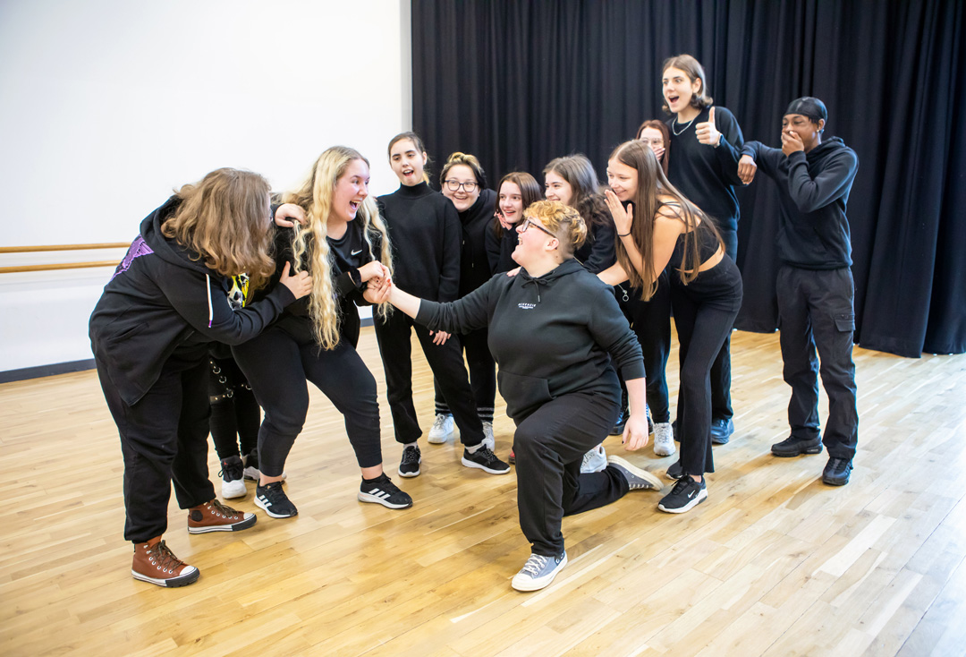Performing arts students gathered in a studio rehearsal, reacting enthusiastically during a group improvisation exercise.