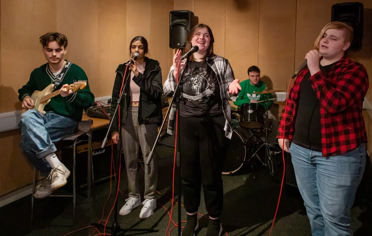A group of five students rehearsing, featuring a guitarist, two vocalists, a bassist, and a drummer in a music studio. A group of five students rehearsing, featuring a guitarist, two vocalists, a bassist, and a drummer in a music studio.