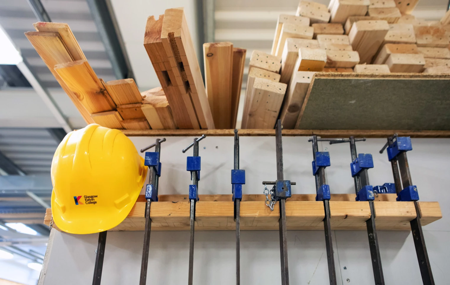 Woodworking equipment including clamps, timber planks, and a bright yellow safety helmet neatly arranged on a storage rack in a joinery workshop. Woodworking equipment including clamps, timber planks, and a bright yellow safety helmet neatly arranged on a storage rack in a joinery workshop.
