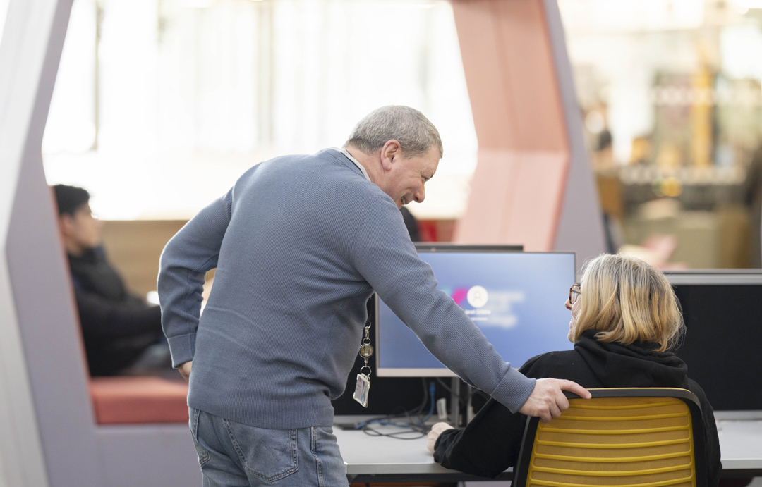 Staff member at Glasgow Kelvin College offering friendly support to a student seated at a computer in the library or learning centre.
