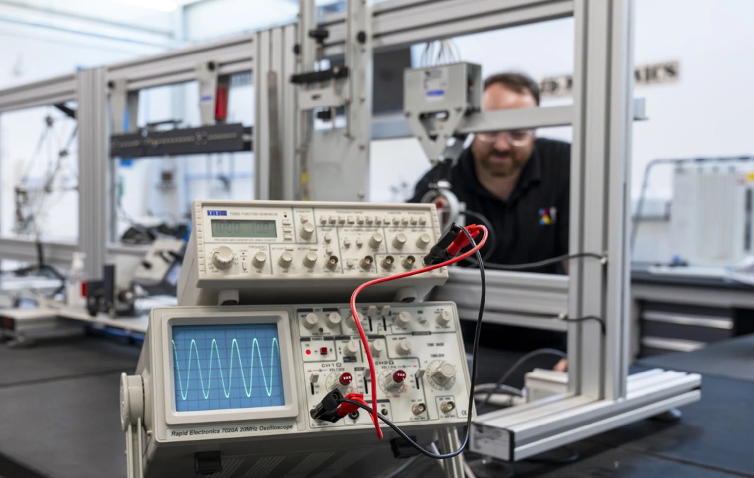 Close-up of an oscilloscope displaying a waveform, connected to a function generator in a laboratory, with a lecturer working in the background. Close-up of an oscilloscope displaying a waveform, connected to a function generator in a laboratory, with a lecturer working in the background.