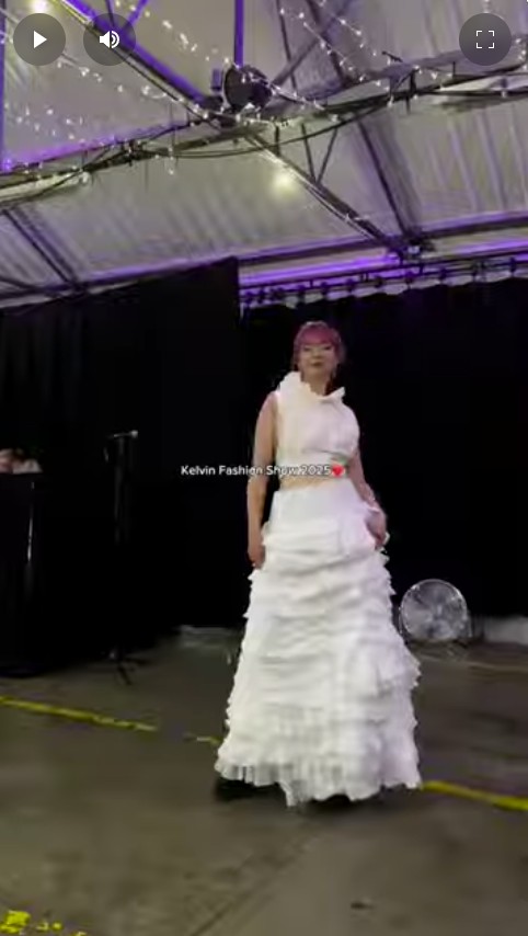 Student model wearing a ruffled white dress walking the runway at the Kelvin Fashion Show 2025, under purple lighting and string lights.