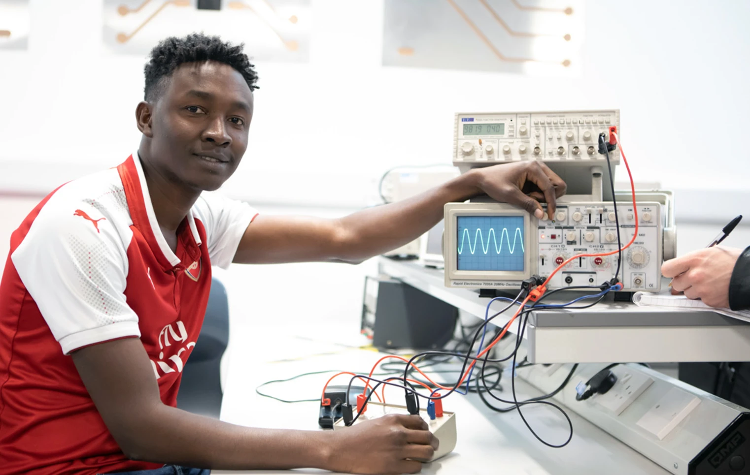 A student in a red football jersey adjusting an oscilloscope in an electronics lab, looking at the camera with a confident expression. A student in a red football jersey adjusting an oscilloscope in an electronics lab, looking at the camera with a confident expression.