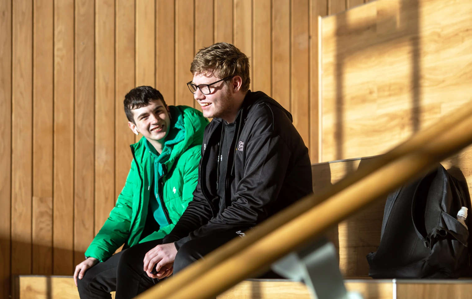 Two students sitting on tiered wooden seating at Springburn campus, chatting and enjoying the sunlight. Two students sitting on tiered wooden seating at Springburn campus, chatting and enjoying the sunlight.