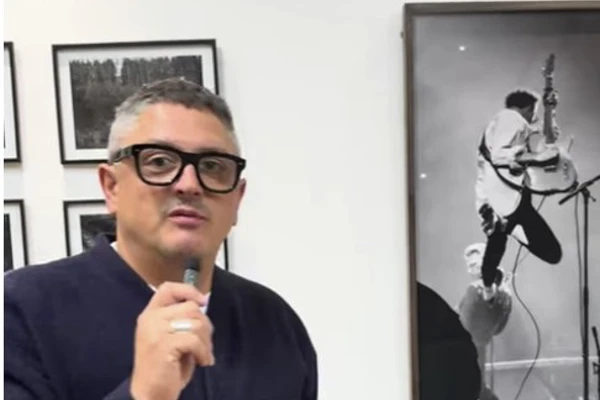 Photographer Mark Cameron speaking at an exhibition, standing in front of a wall of framed black and white portraits, including an action shot of a guitarist. Photographer Mark Cameron speaking at an exhibition, standing in front of a wall of framed black and white portraits, including an action shot of a guitarist.