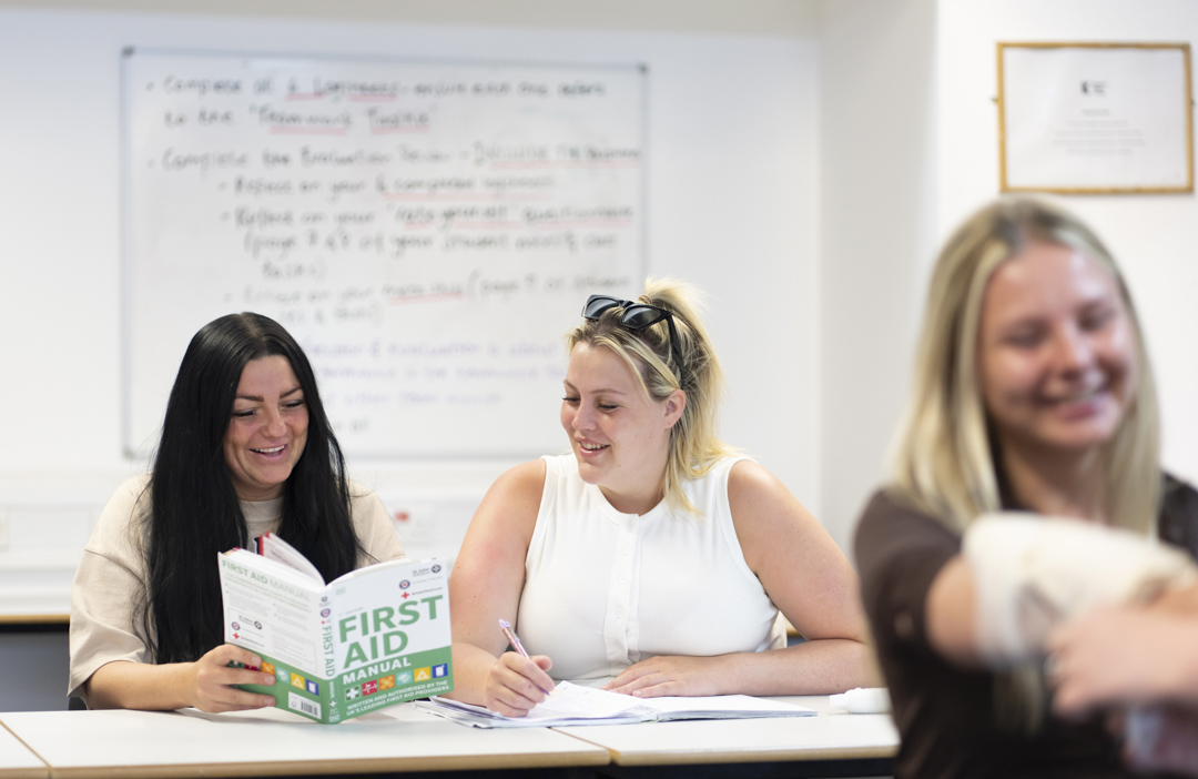 Two students sit at a classroom desk, one reading a first aid manual and the other taking notes, both focused and ready to learn with a whiteboard full of notes behind them.
