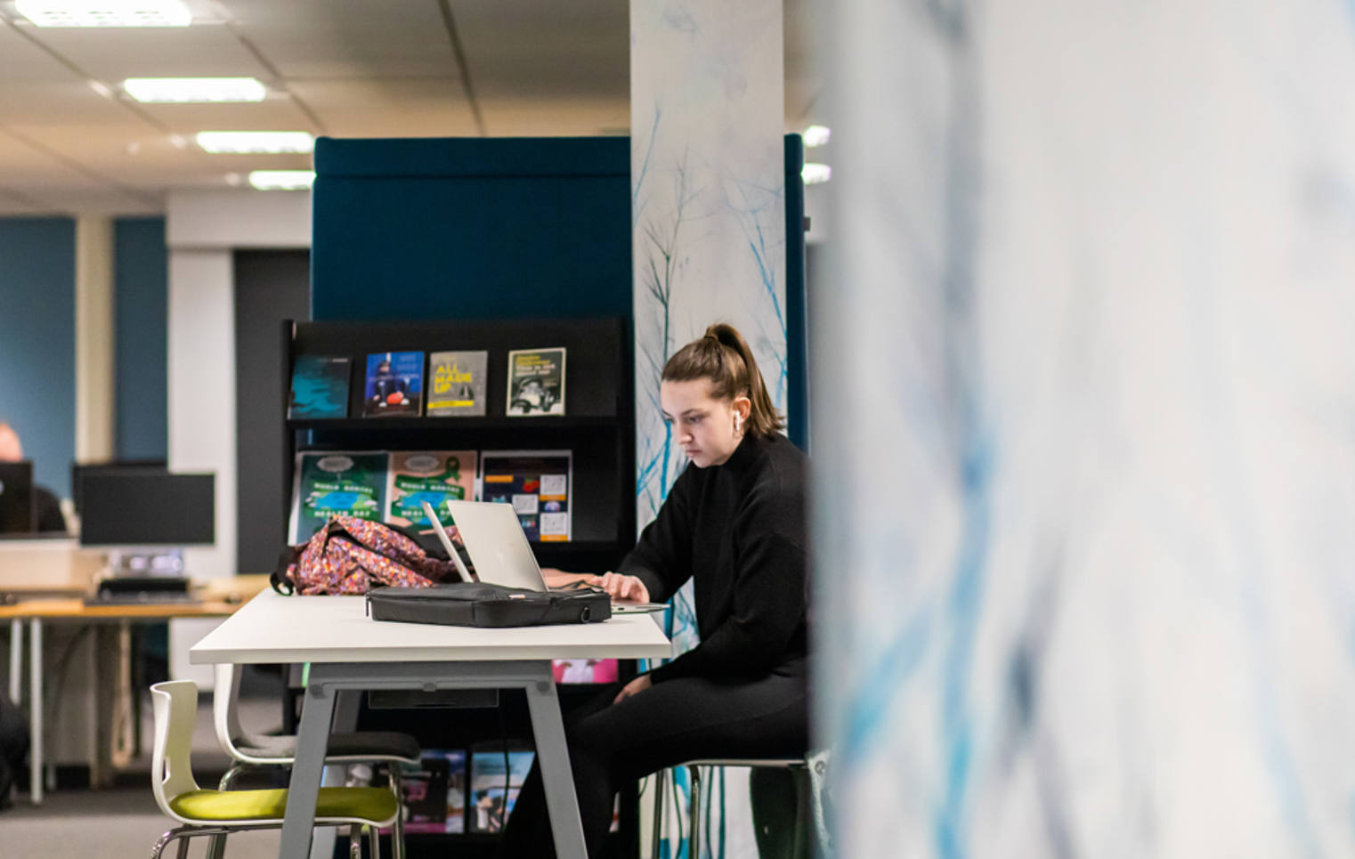 Student focused on studying with two laptops at a desk in front of a reference bookshelf. Student focused on studying with two laptops at a desk in front of a reference bookshelf.