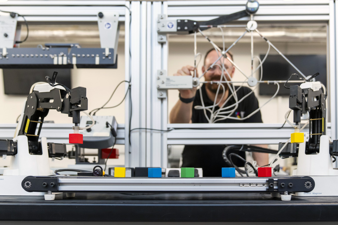 A lecturer working behind an advanced robotics training system, adjusting wiring while robotic arms manipulate colored blocks.