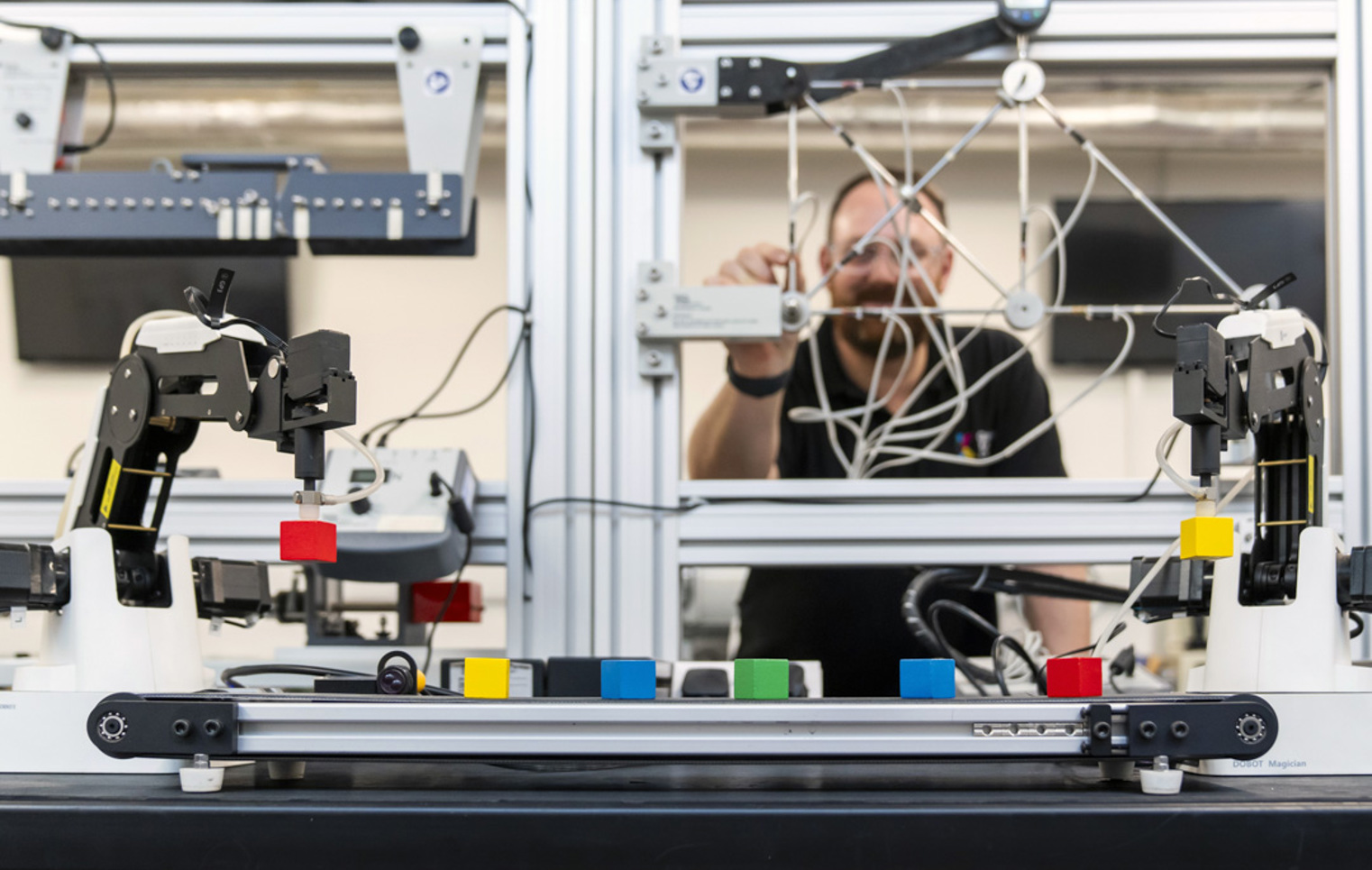 A lecturer working behind an advanced robotics training system, adjusting wiring while robotic arms manipulate colored blocks.